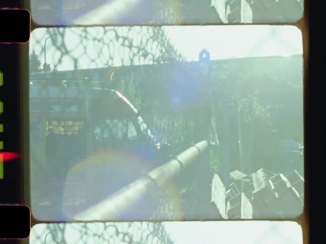 A train passes near a wire fence with a yellow van parked behind it as bright sunlight floods the scene