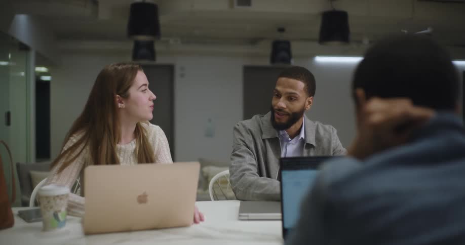 The three coworkers are sitting at the table with their laptops and they are talking to each other