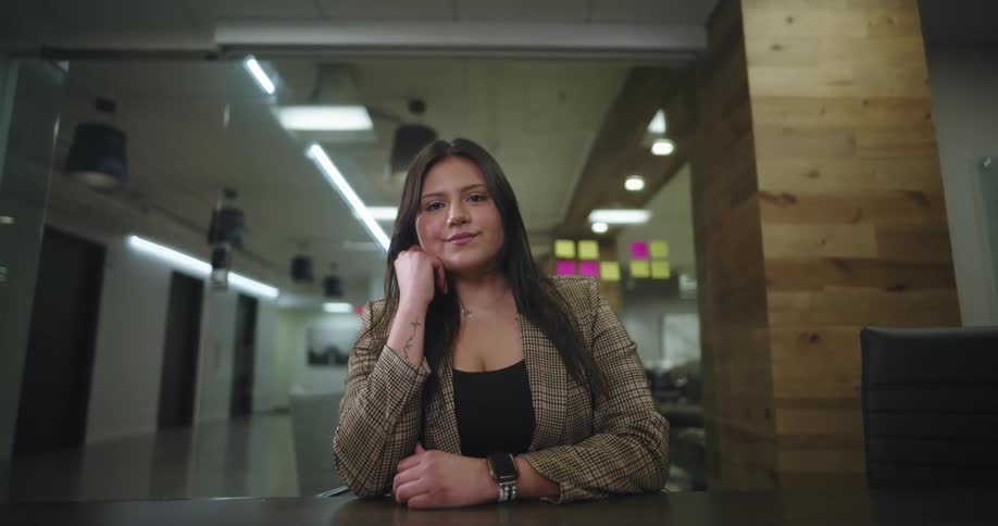A woman with long hair is seated at a table looking forward in a modern office environment featuring a glass wall and wooden accents