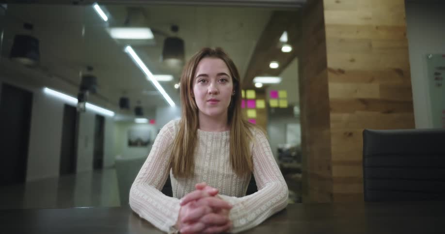 A woman with blond hair sits at a desk in a modern office facing forward with hands clasped as she looks ahead surrounded by glass walls warm lighting and colorful sticky notes in the background