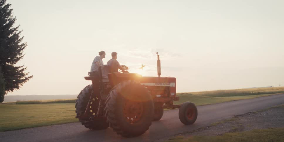 Two individuals are riding on a red tractor moving along a paved road during a bright possibly early morning or late afternoon
