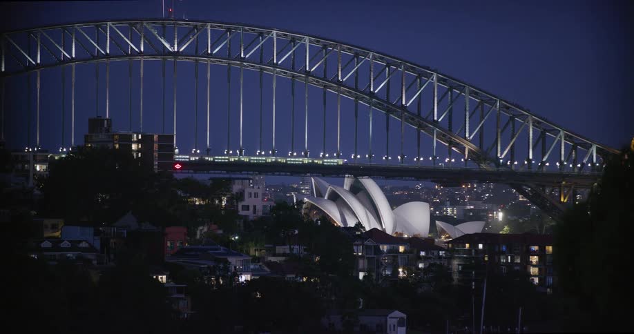 A nighttime shot of the sydney harbour bridges steel arch structure illuminated against a dark blue sky frames the sydney opera house in the distance