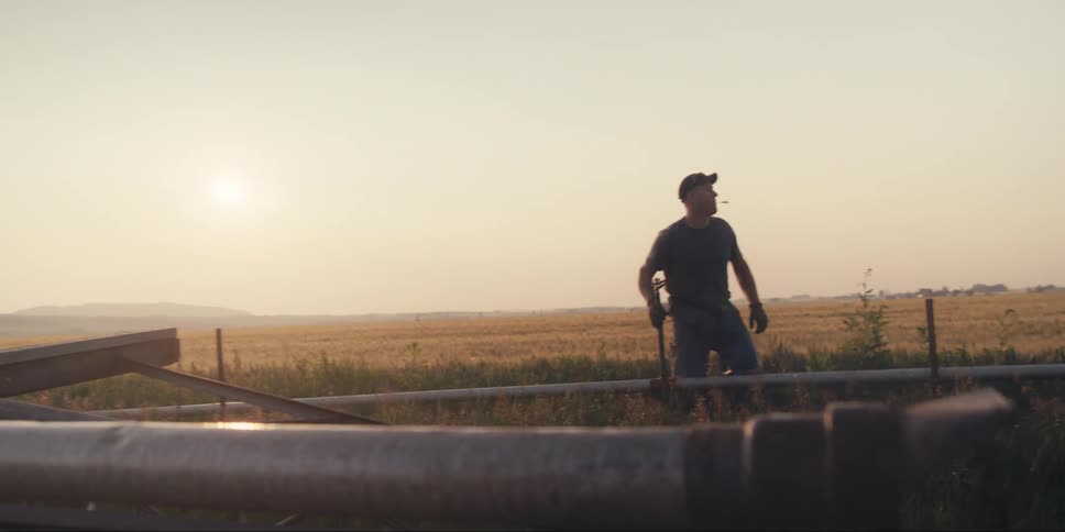 A farmer in a rural field silhouetted against the bright sunlight bends to lift a section of irrigation pipe