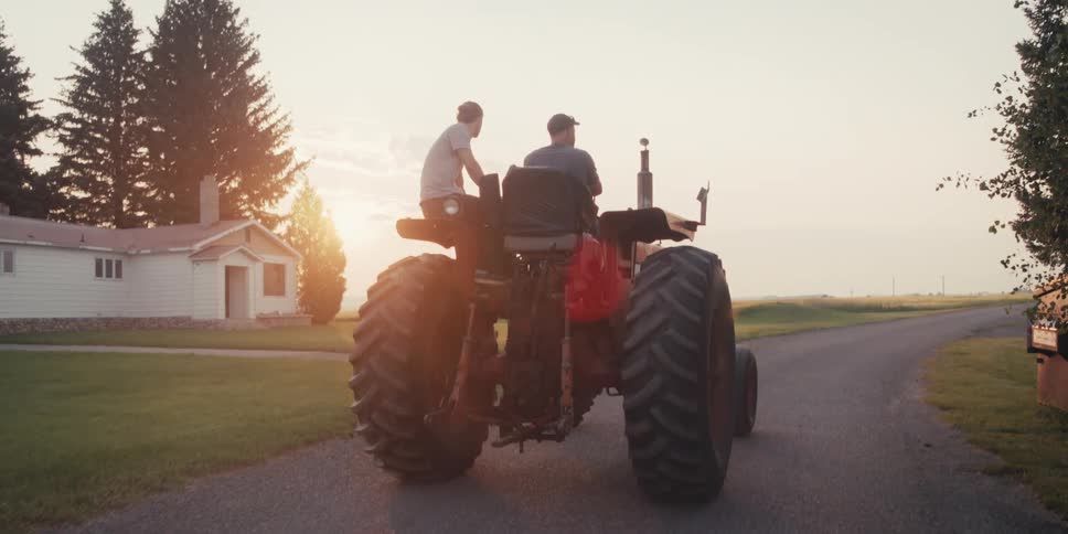 The two men are sitting on the tractor as it is being driven down the road by the older man and they go passed a house and a group of trees on either side of the road