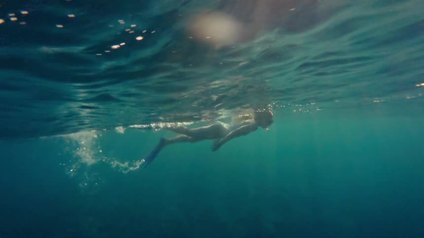 A young woman with lightcolored hair swims underwater in a light colored swimsuit and blue flippers with sunlight filtering through the water