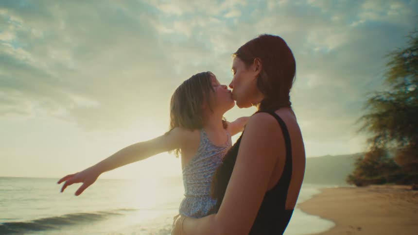 A mother holds and kisses her little daughter on the beach at sunset