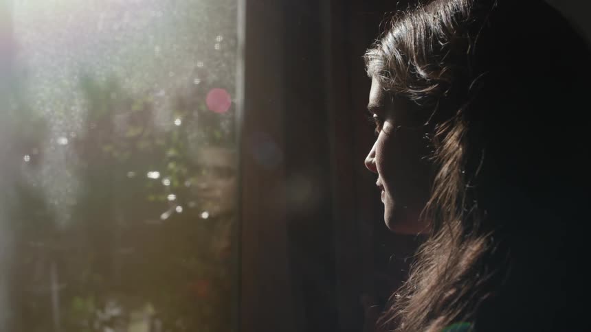 A woman stares out an old window as the sun shines