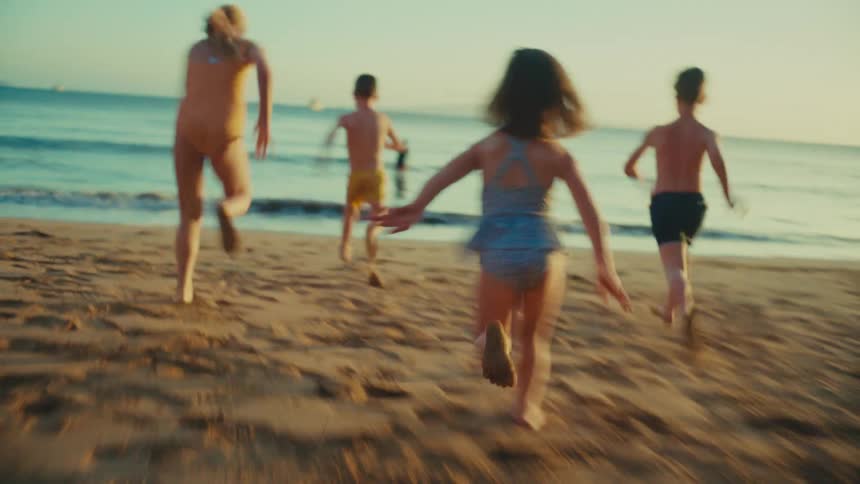 A group of children wearing swimsuits running towards a woman standing in the shallow water of a beach at sunset