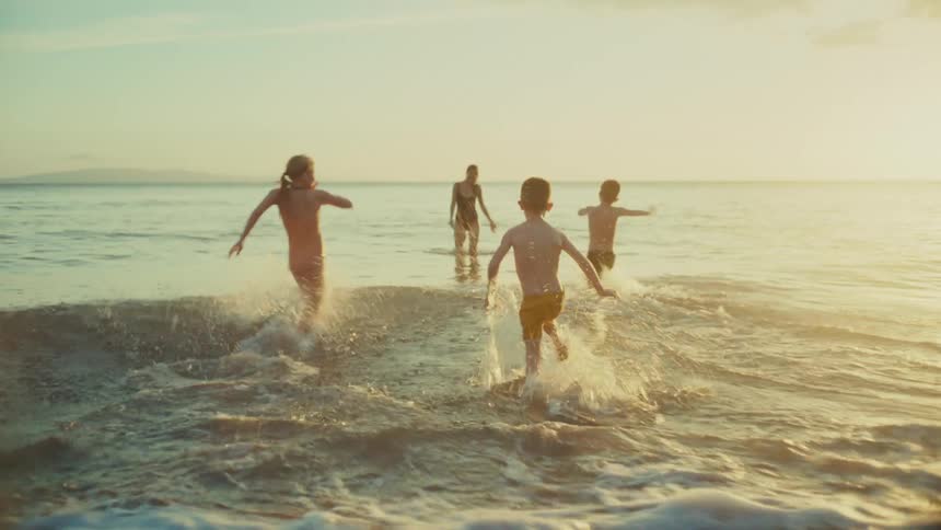 Children in swimwear run joyfully along the beach at sunset heading toward their mother and the ocean