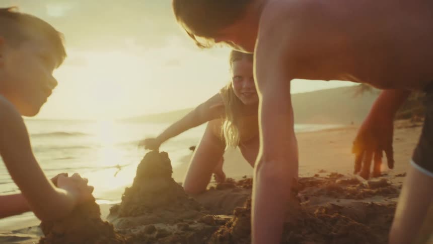 Three children play in the sand on a beach at sunset building a sandcastle
