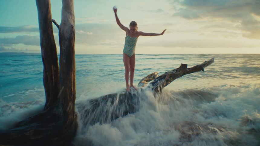 A young girl balances with outstretched arms on a large piece of driftwood in the ocean with the horizon and a cloudy sky in the background