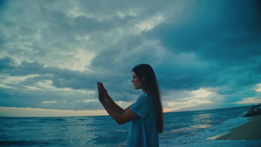 A woman with long dark hair stands on a beach near the ocean holding a phone against a cloudy sky