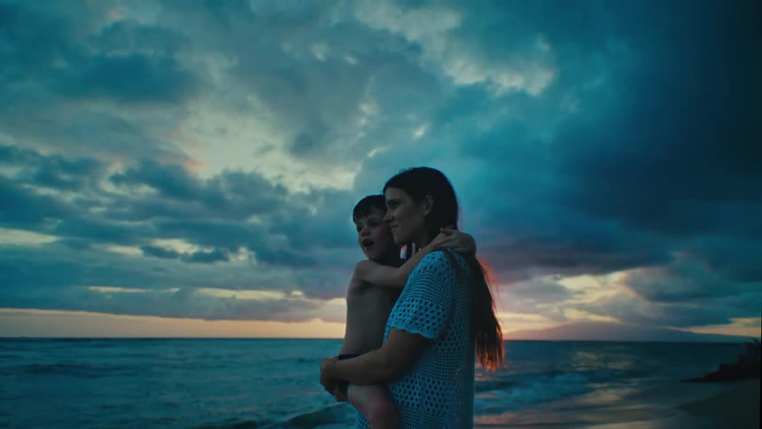 The mother is holding her son on the beach beside the ocean and waves crashing