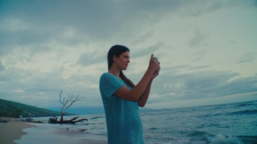 A woman with long dark hair wearing a blue patterned top holds a smartphone up to her face taking photos on a beach with a dramatic sky and ocean in the background