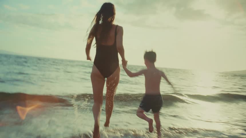 A mother and her son wearing swimwear run joyfully along the beach at sunset with the coastline in the background