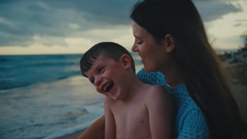A mother sits on the beach at sunset with her son in her lap sharing kisses and a joyful conversation by the coast