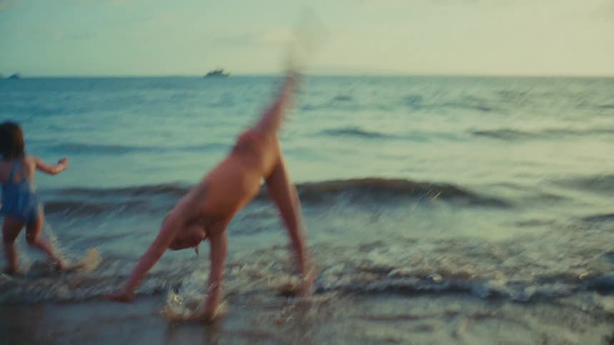 Girl cartwheeling in shallow waves with her younger siblings playing in the water behind her