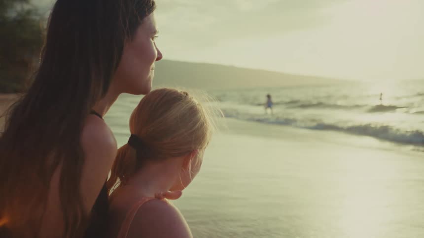 A mother sits on a beach with her daughter on her lap kissing her head under the soft sunlight of either a sunset or a morning