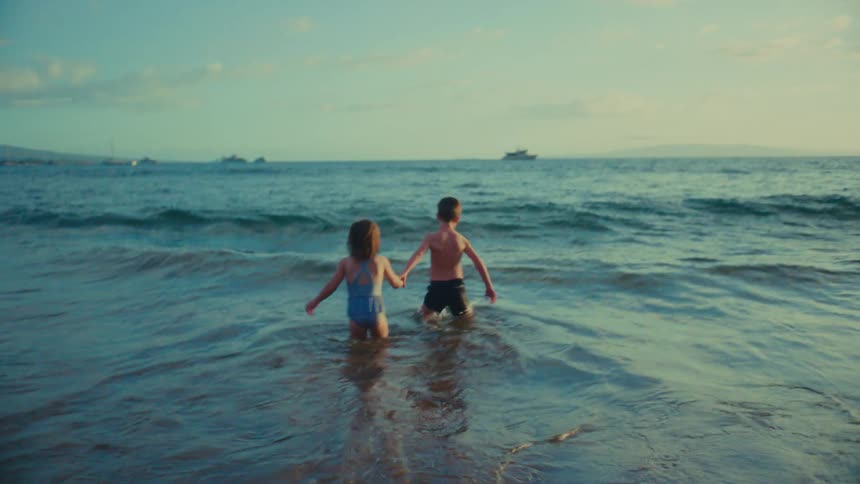 Two children a girl in a blue swimsuit and a boy in dark shorts stand waist deep in the ocean facing away from the viewer with boats visible in the distance