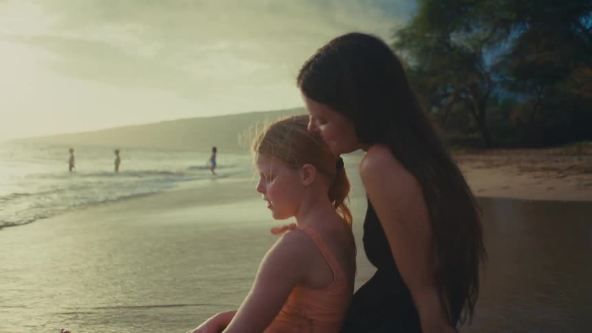 A mother and daughter sit on a beach at sunset with the woman embracing the child and other people visible in the distance