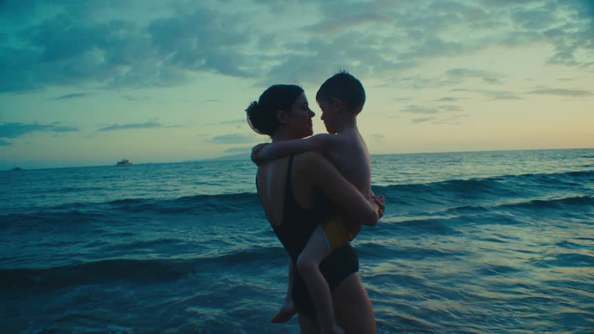 Mother kissing young son while he points toward ocean on tropical beach at sunset