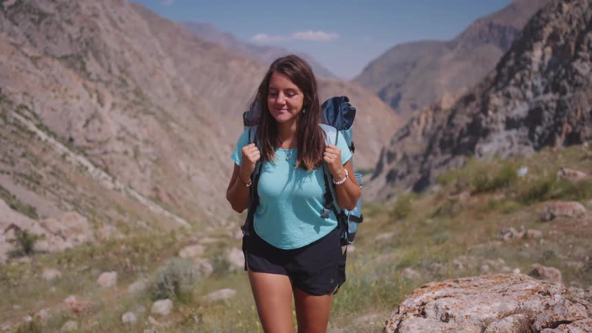 A young woman with long brown hair wearing a blue shirt dark shorts and a backpack walks through a rocky mountainous landscape