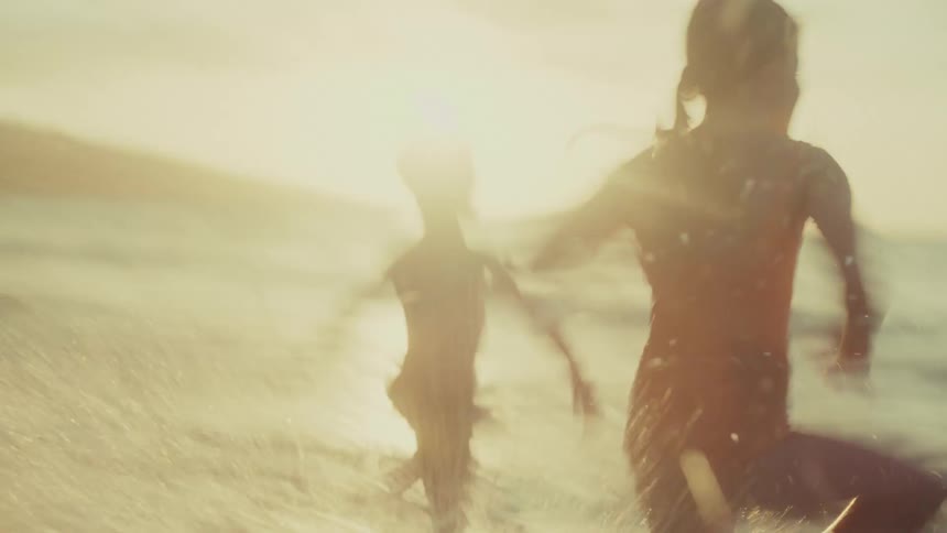 A boy and a girl joyfully run across a sandy beach toward the ocean with the sunset casting a warm glow