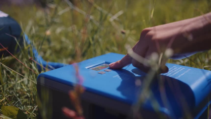 A persons finger presses a button on a rectangular blue machine placed in the grass