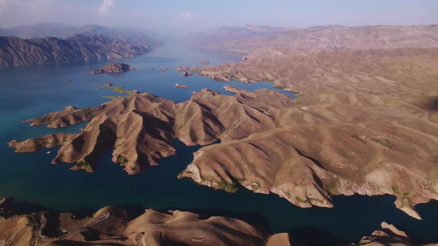 A sweeping overhead view reveals winding desert hills jutting from a sprawling lake with scattered islands under a clear sky capturing the rugged textures vast contours arid grandeur and shimmering waters in warm sunlight