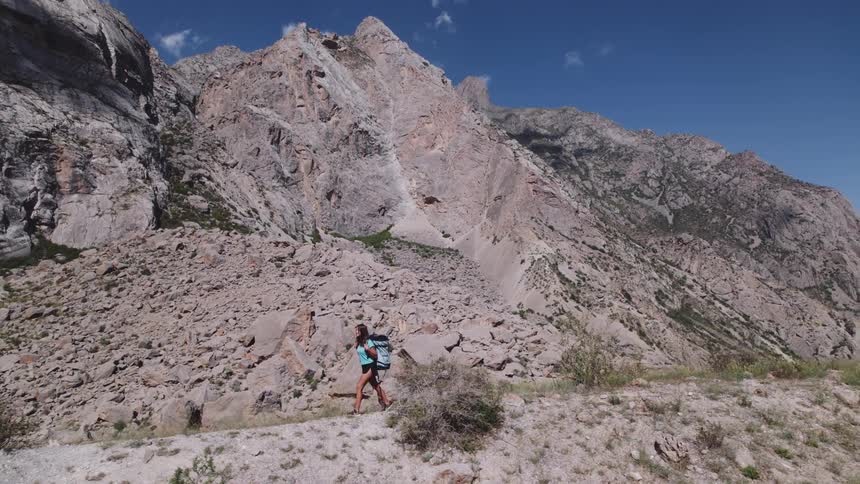 A woman with a backpack hikes along a rocky trail at the base of a large rugged mountain under a clear blue sky
