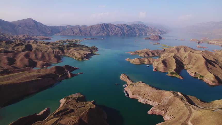 Aerial view captures a vast lake surrounded by rolling dry hills and rugged mountain ranges in the distance with winding shorelines and calm waters extending beneath a clear blue sky