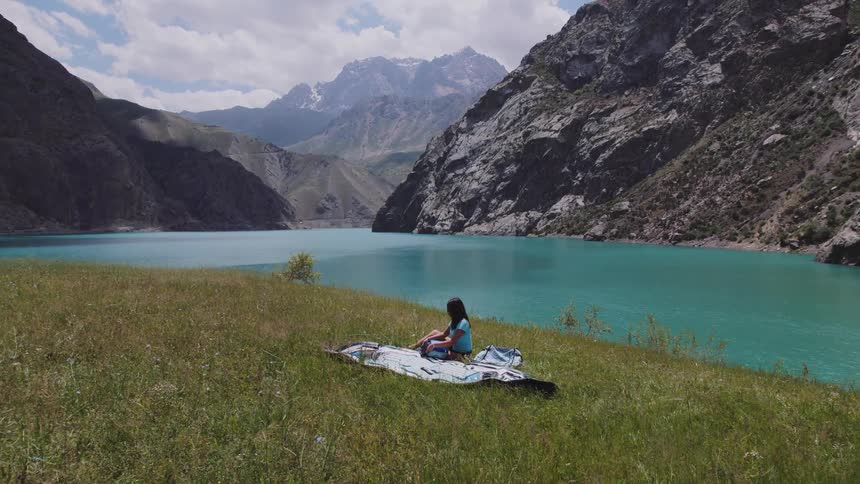 A woman sits and picks up her thermos in a field surrounded by mountains
