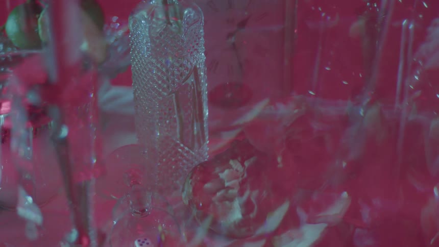 A closeup view shows a glass vase a bottle and cups arranged on a pink tablecloth with roses slightly distorted and out of focus