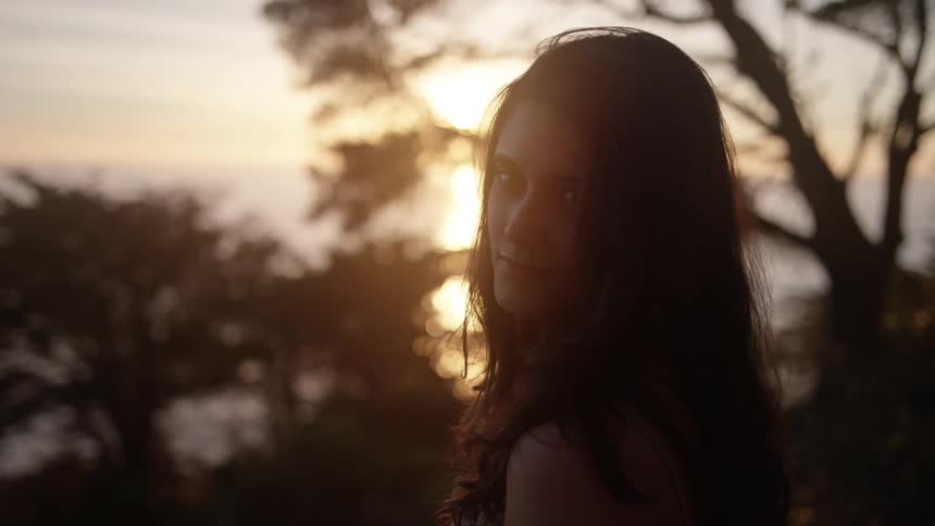 A woman standing by a body of water and trees at sunset