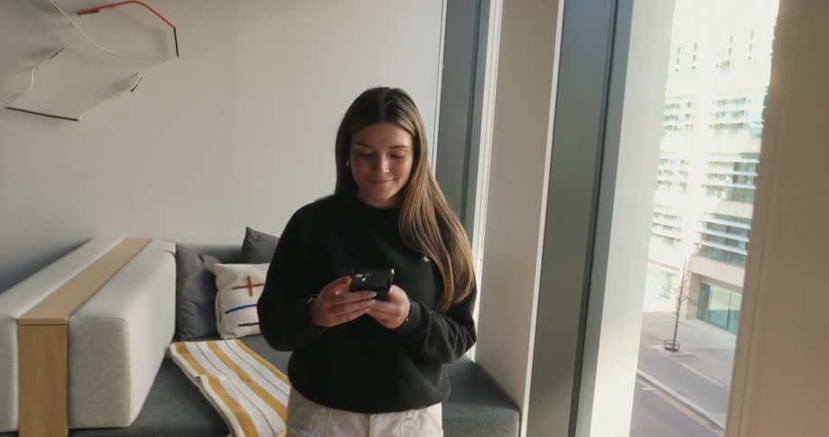 In a twisting motion the view zooms into a woman standing in a modern office lounge as she texts on her phone near tall windows overlooking a city street and surrounding buildings