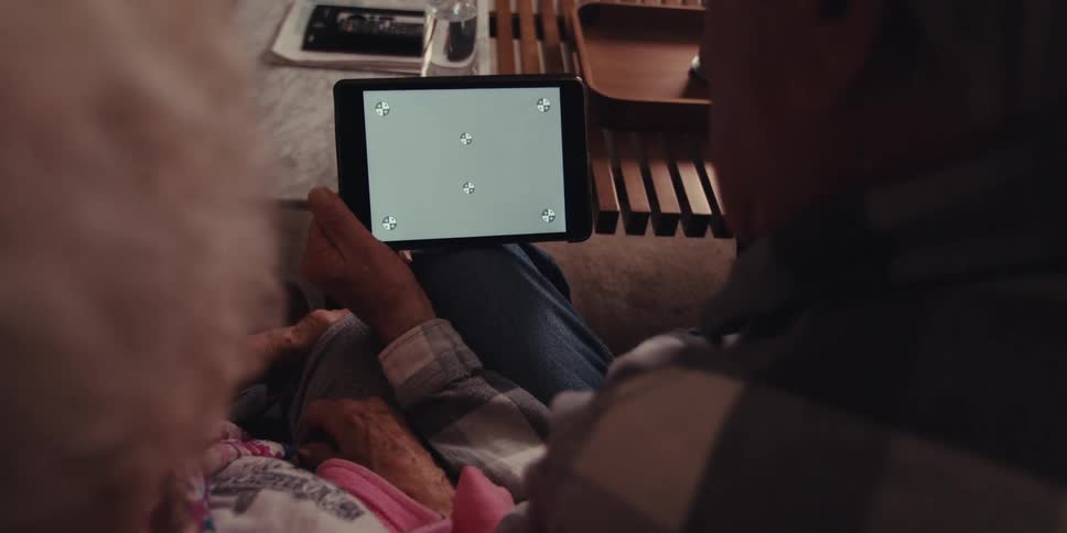 Elderly man with a plaid shirt holds a tablet and woman looks down at the screen sitting on the couch in the living room