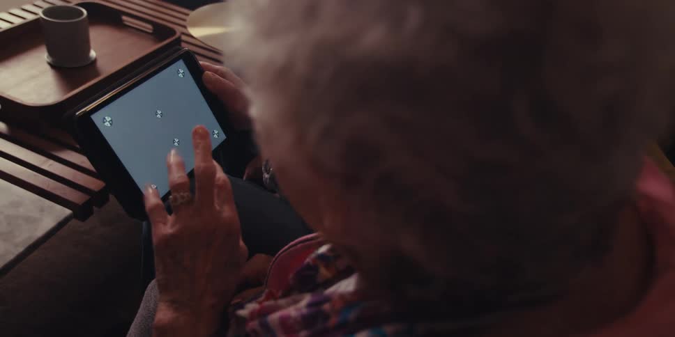 An elderly man and woman with gray hair use a tablet while sitting on a couch in the living room