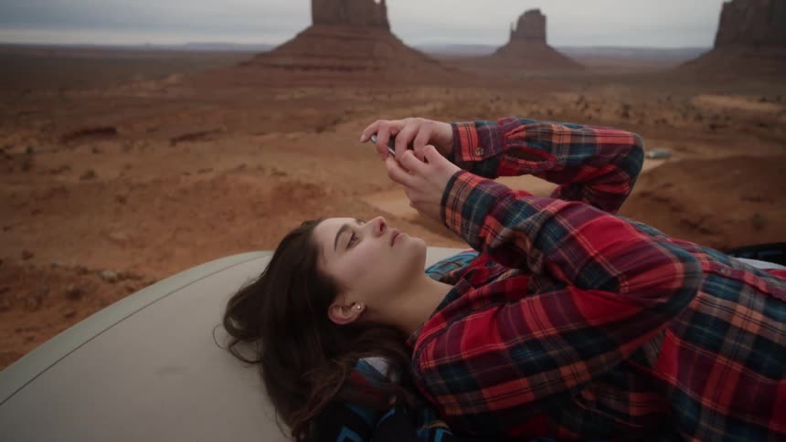 Woman lays on blanket on a car and plays with her phone