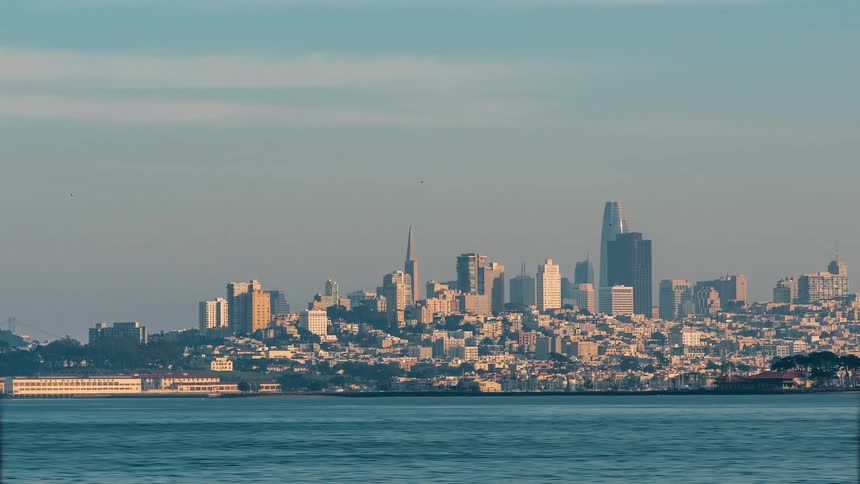 The sunlit cityscape of san francisco rises from the edge of the bay with its iconic skyline silhouetted against a hazy sky