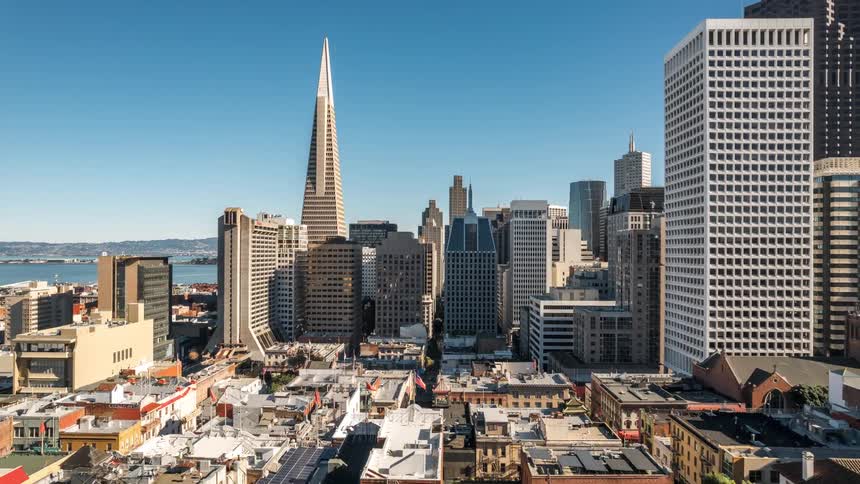 A cityscape view showcases the iconic transamerica pyramid among other tall buildings against a clear blue sky with a glimpse of the bay in the distance