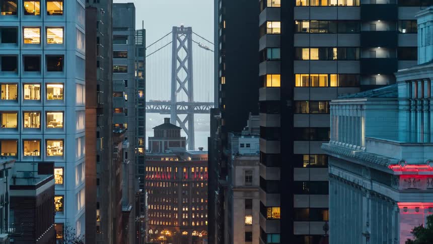 A view through a narrow gap between tall buildings reveals the bay bridge in the distance with lit windows in the surrounding structures