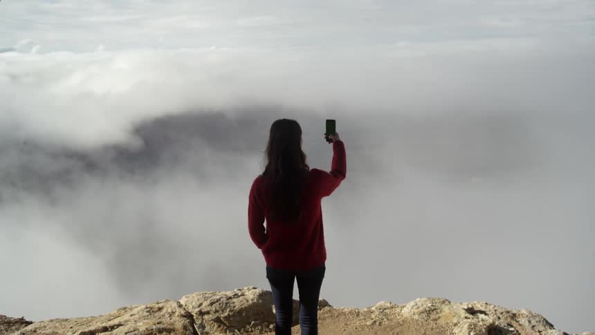 A woman stands near the edge of a cliff and takes a photograph