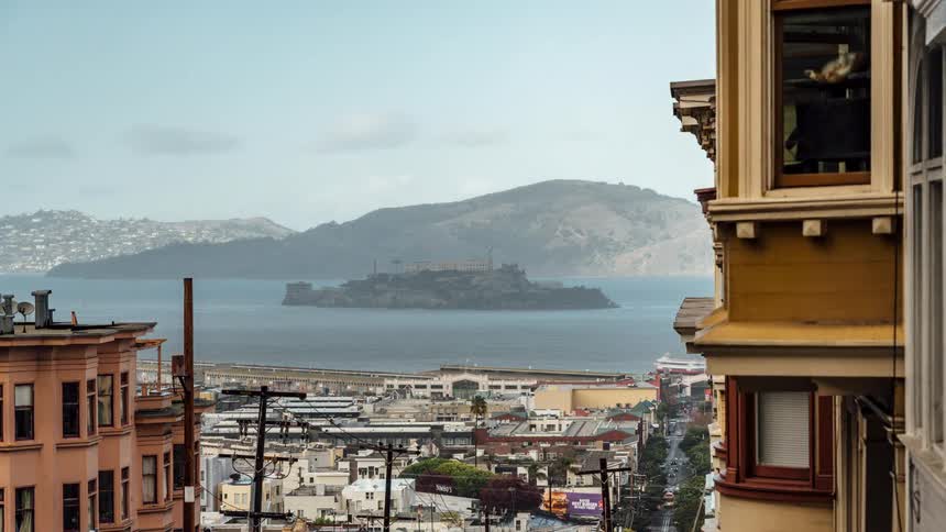 Alcatraz island sits in the bay viewed from between buildings on a hilly san francisco street
