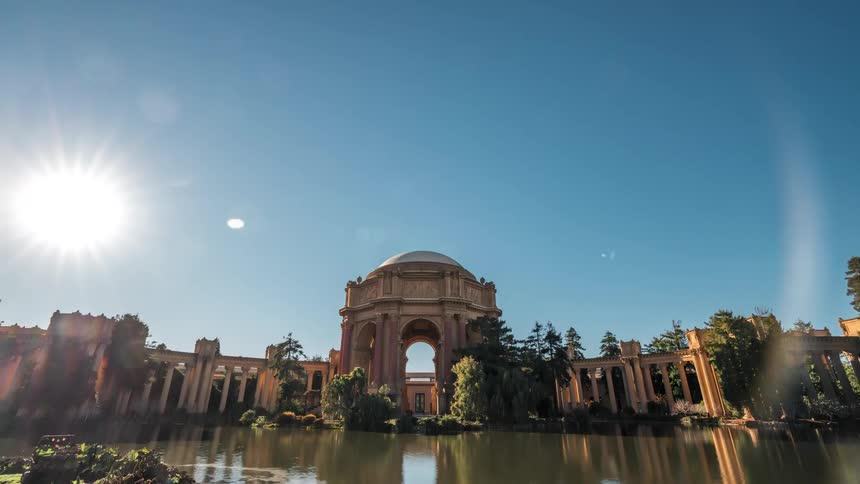 The palace of fine arts with its distinctive dome and colonnades is reflected in a calm lagoon under a bright sun with people scattered along the foregrounds edge