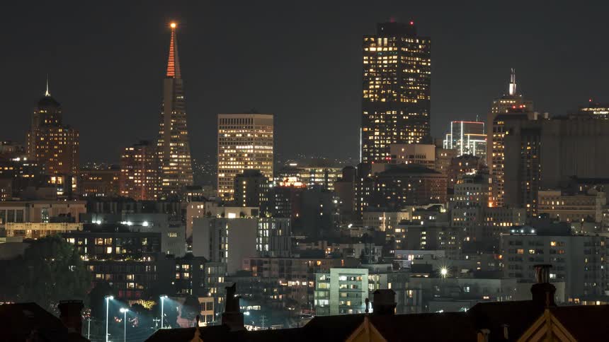 A nighttime cityscape from a high vantage point showcasing a dense urban area with numerous illuminated buildings including iconic skyscrapers against a dark sky