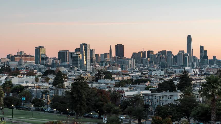 The san francisco skyline at dusk with buildings silhouetted against a soft pastel colored sky seen from an elevated perspective with trees and park greenery in the foreground