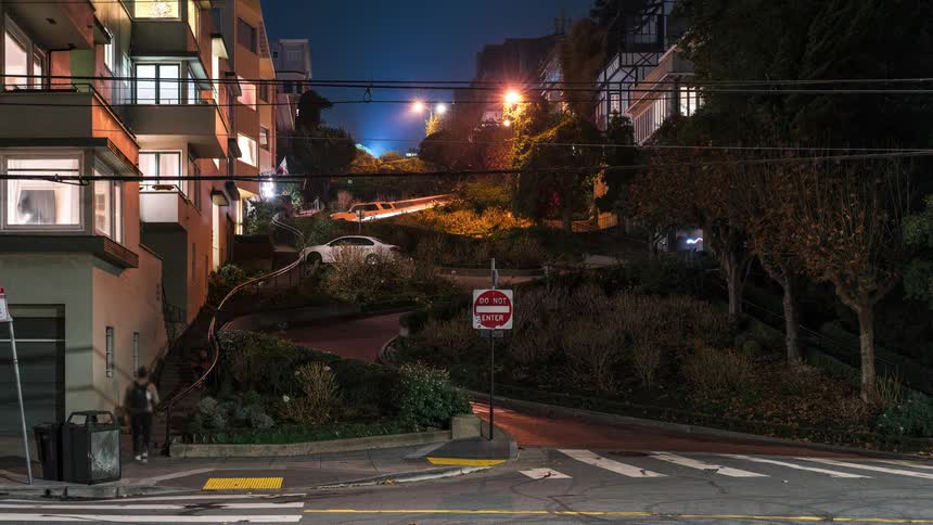A winding street ascends a hill at night illuminated by streetlights and the warm glow from apartment windows with a do not enter sign prominently displayed at the intersection