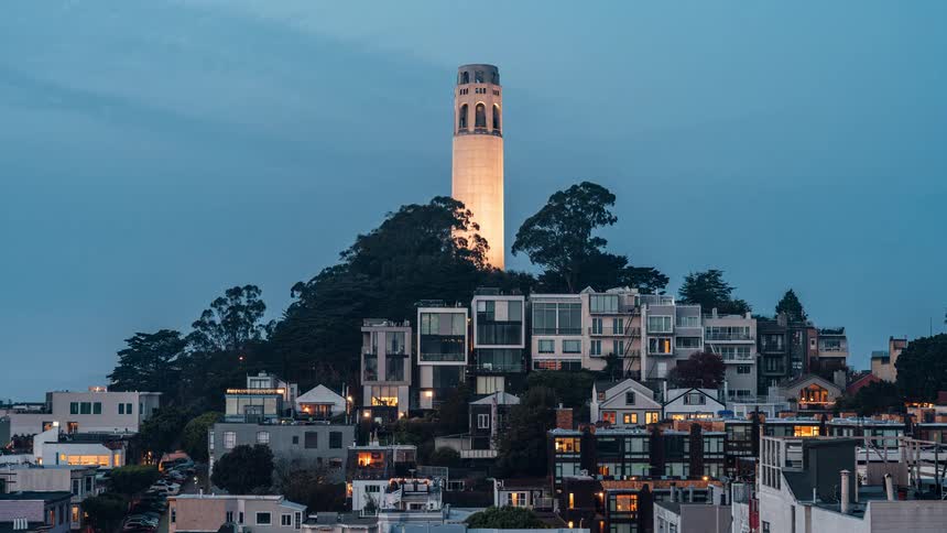 A tall light colored tower stands prominently atop a hill amidst a neighborhood of various residential buildings under a soft blue tinted sky