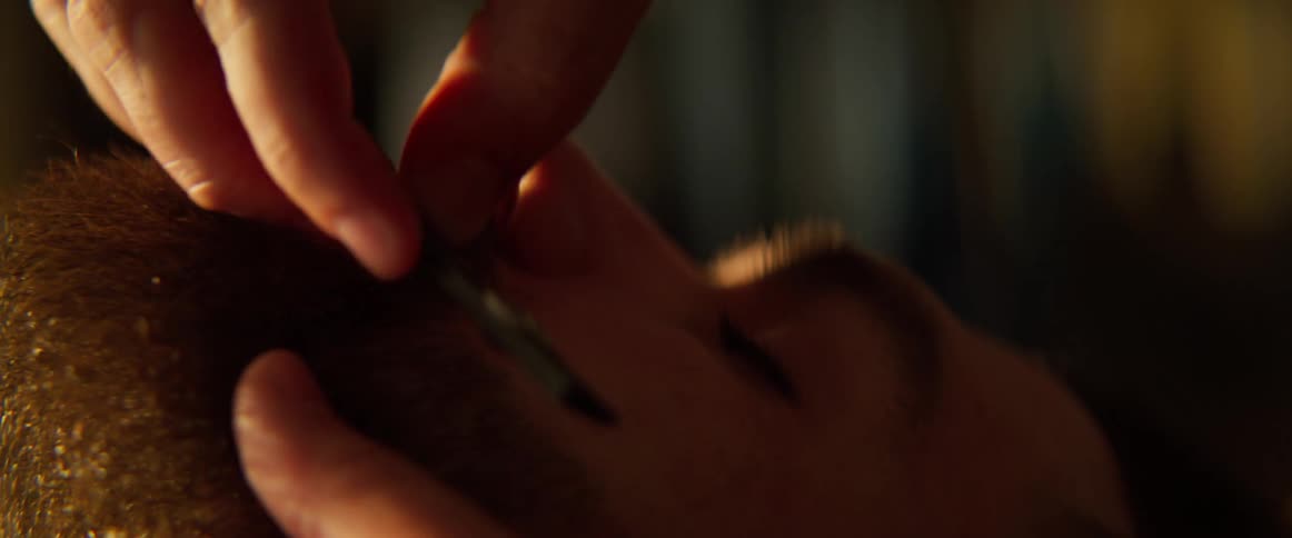 A persons hand using a razor to shave the beard of a man who has his eyes closed