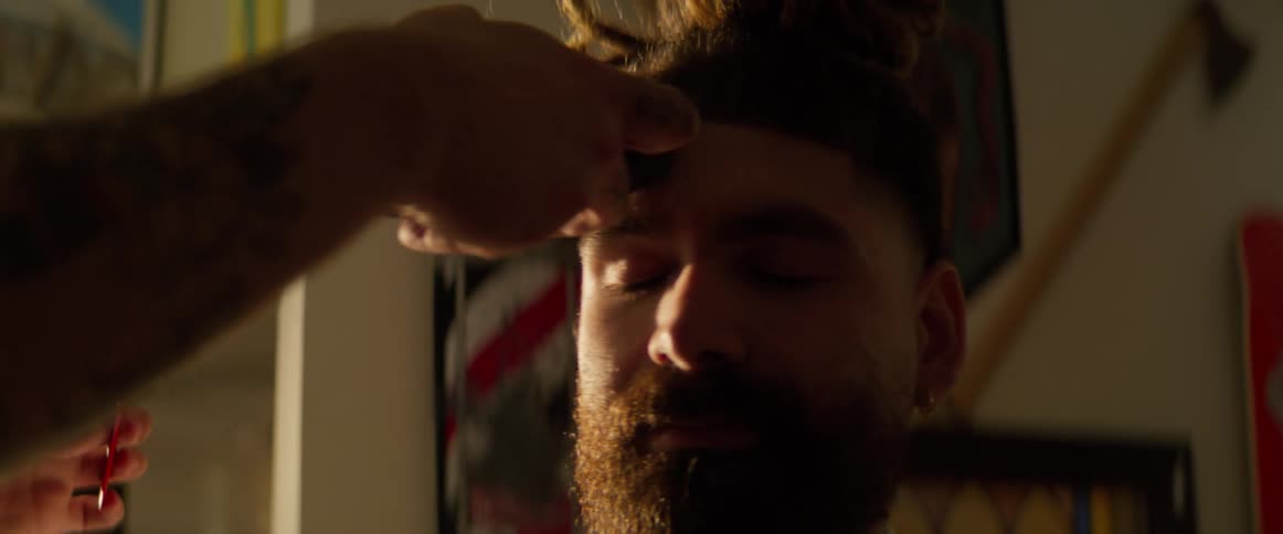 A closeup of a man with a bun as a barber trims and combs his hair with a cape draped around him in a dimly lit barber shop